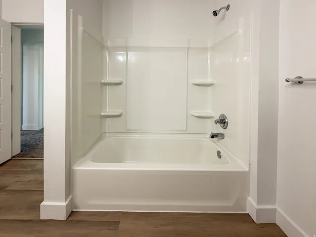 White bath and shower combination with chrome fixtures and integrated shelving, set against wood-finish flooring