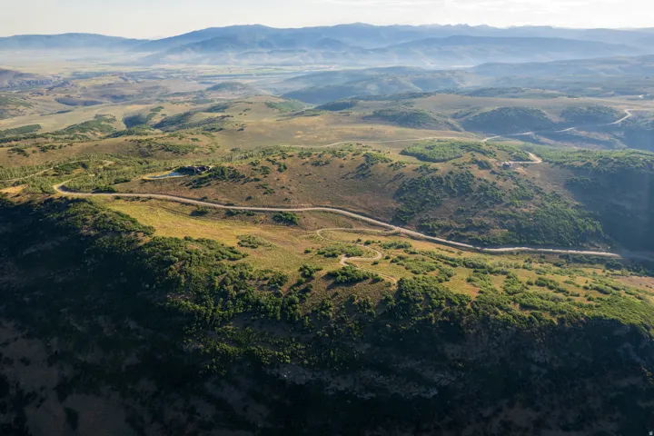 Expansive mountain landscape featuring varied terrain with distant peaks, a winding road, and scattered evergreen and deciduous trees