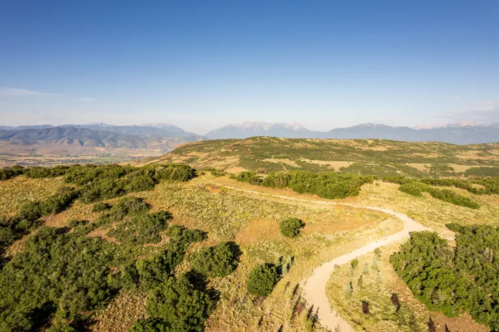 Expansive undeveloped land featuring rolling hills, varied green vegetation, a winding dirt road, distant mountain ranges, and a valley landscape