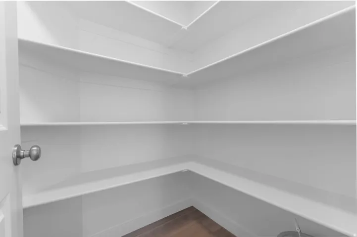 Spacious pantry featuring multiple white built-in shelves, wood-finish flooring, and a white interior door with a satin nickel doorknob