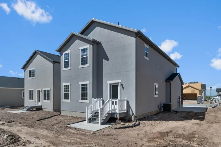 Newly constructed residence featuring a gray stucco exterior, multiple windows with white trim, a rear concrete patio, and white-railed stairs leading to a back entrance