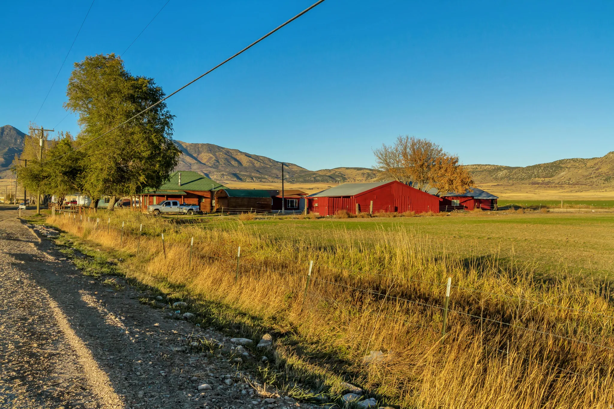 Expansive agricultural property featuring a red barn, additional red outbuilding, a dwelling with a green roof and wood-finish siding, and a vast field with natural grass and distant mountains