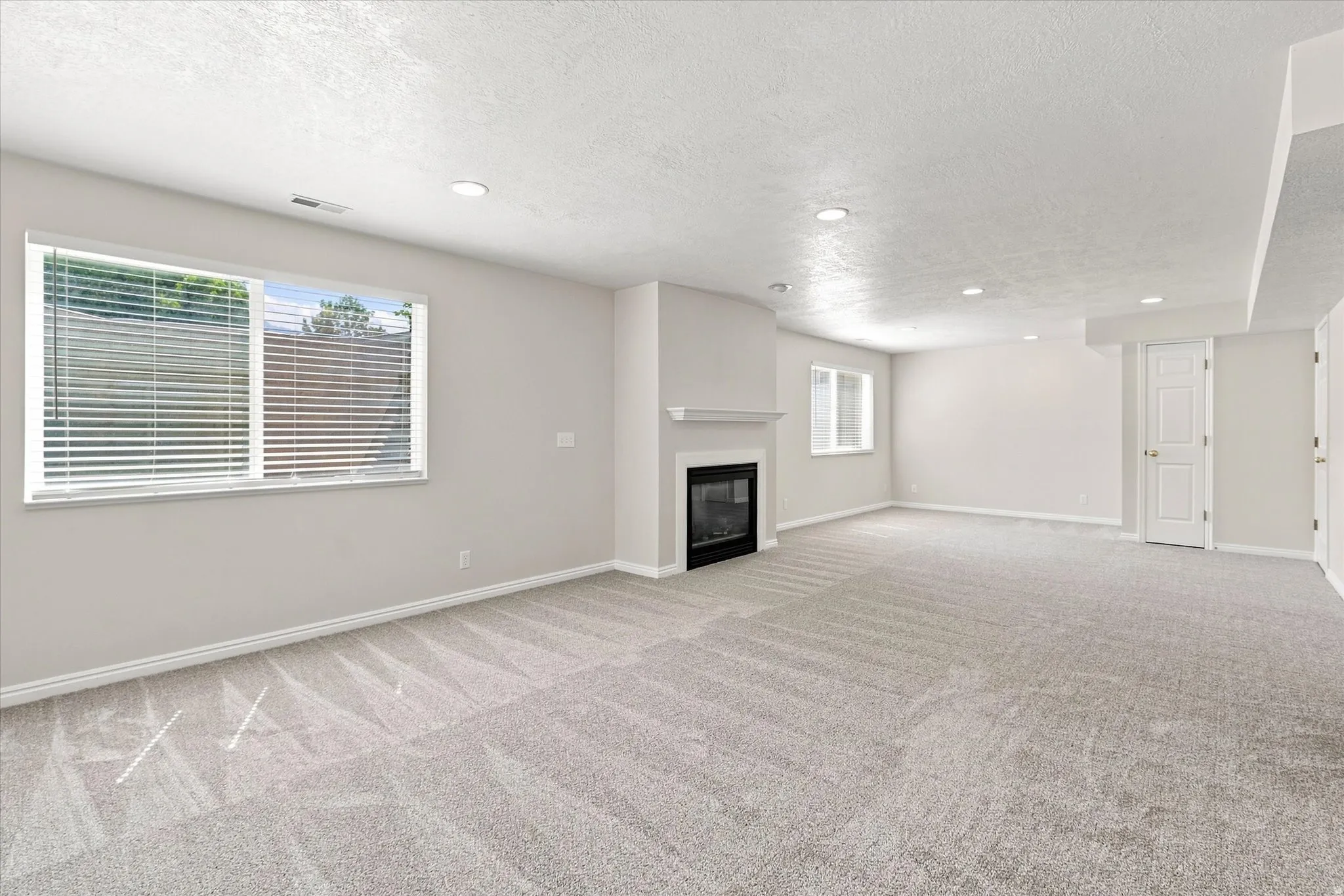 Spacious room with light-colored carpeting, a contemporary fireplace, multiple windows with blinds, recessed lighting, and neutral wall tones