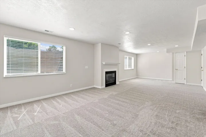 Spacious room with light-colored carpeting, a contemporary fireplace, multiple windows with blinds, recessed lighting, and neutral wall tones