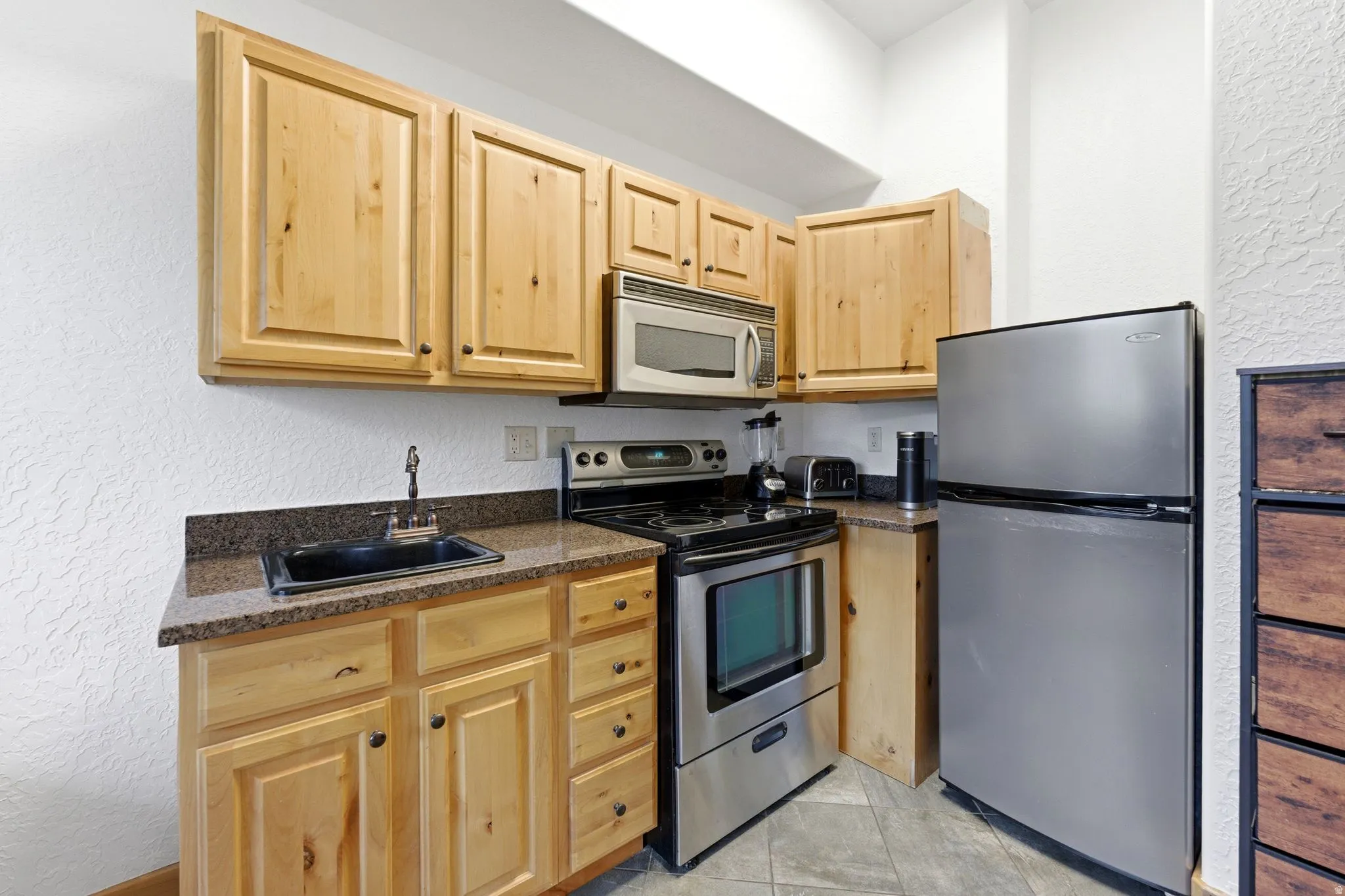 Kitchenette featuring wood cabinetry, dark speckled countertops, and stainless steel appliances including a refrigerator, range, and microwave
