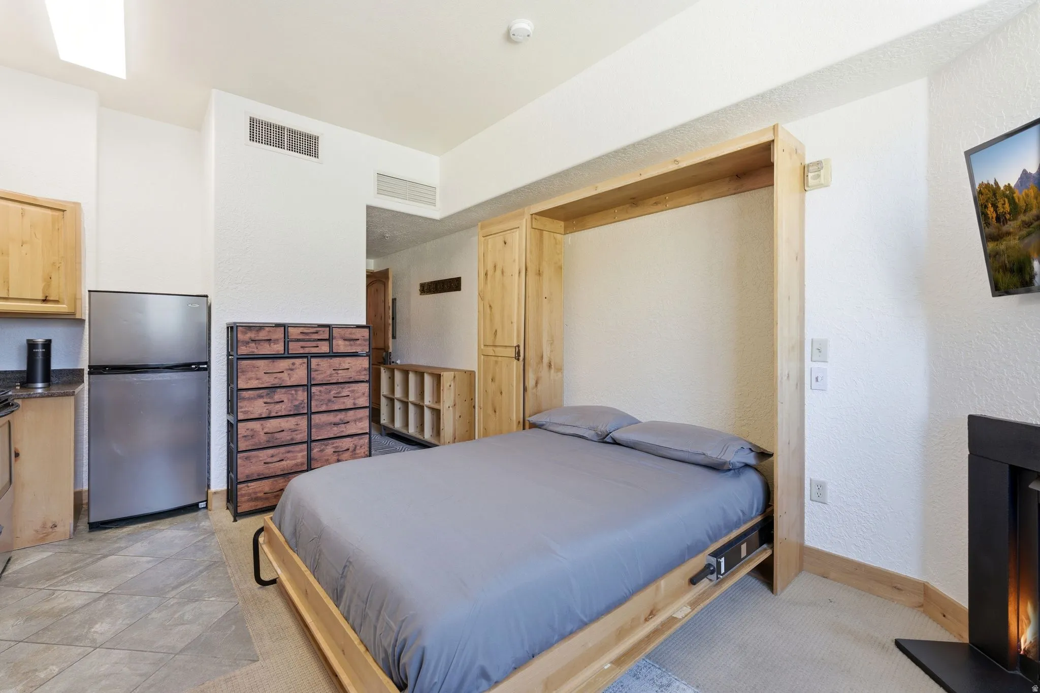 Integrated living space featuring a built-in Murphy bed, a wall-mounted flat-screen television, a stainless steel refrigerator, a compact counter space with a sink, and a black fireplace with a visible flame