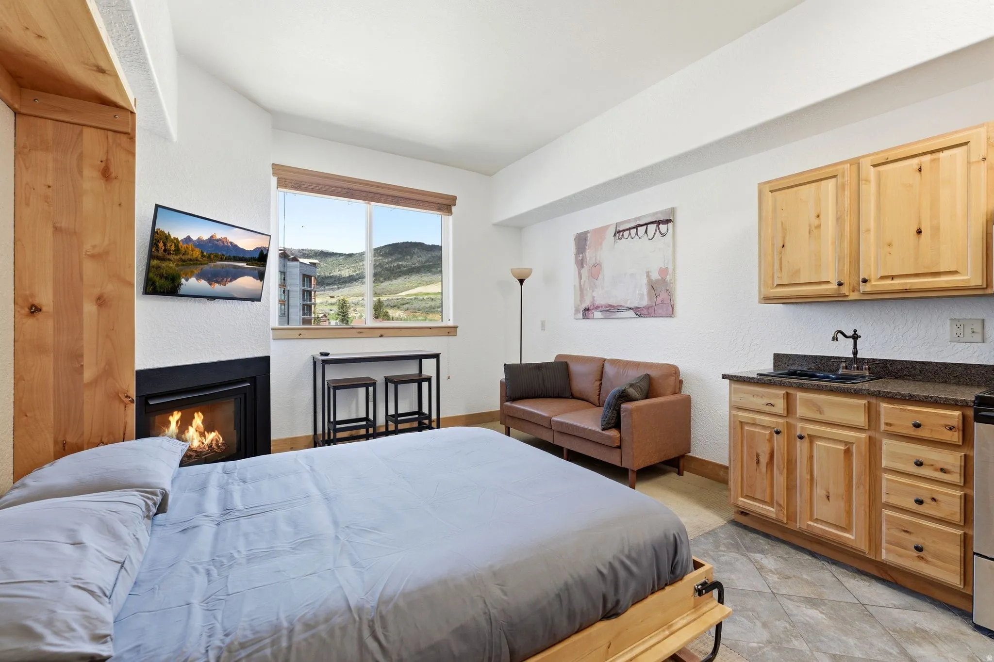 Compact living area featuring a built-in fireplace, a large window showcasing a mountainous landscape, a kitchenette with wood cabinetry and dark countertops, tile flooring, and a dedicated sleeping space