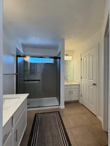 Spacious bathroom featuring a glass-enclosed shower with dark-toned tile, a separate vanity with a white countertop and undermount sink, a second vanity with a white countertop and undermount sink, a large mirror, and tile flooring