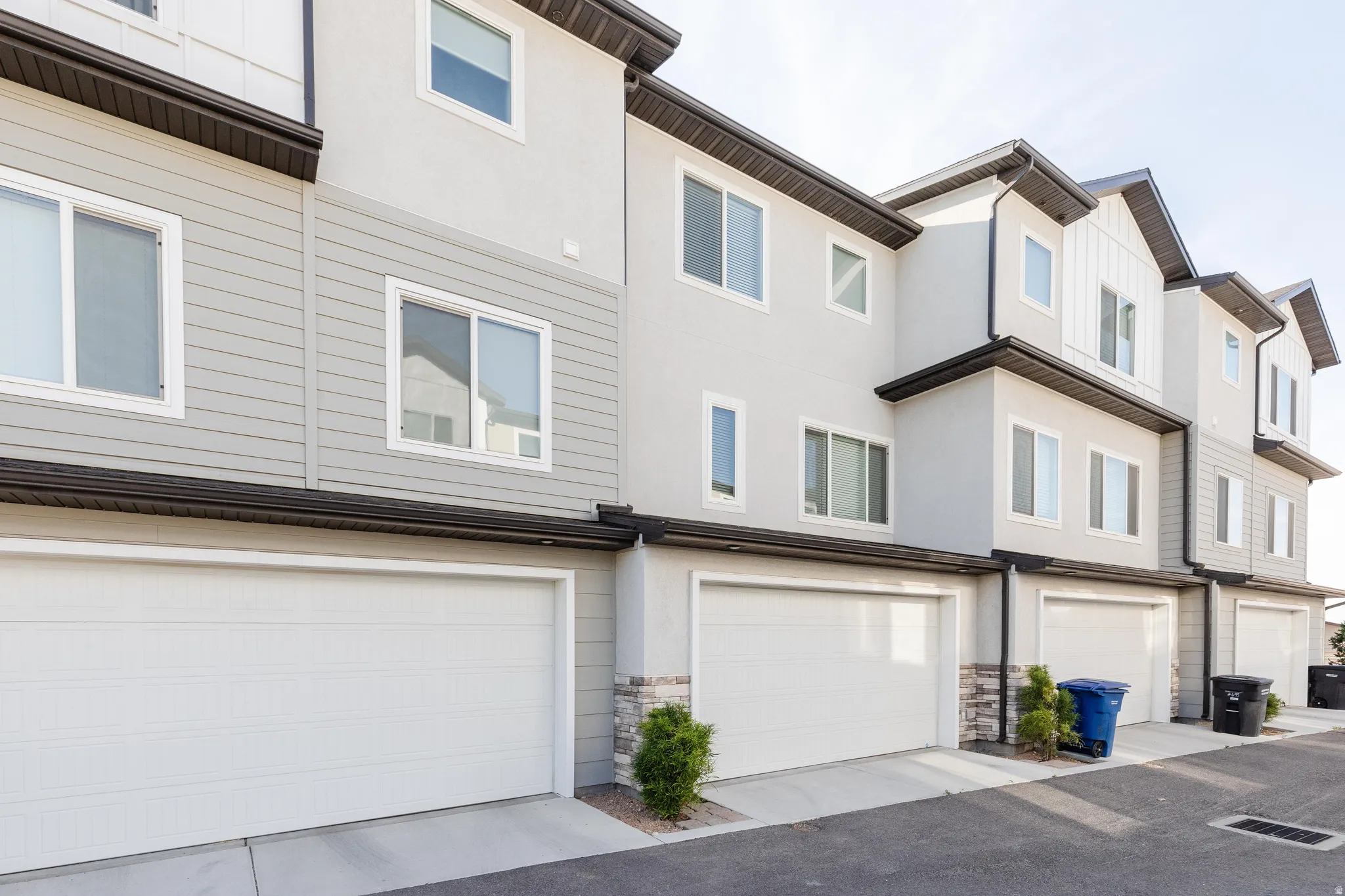 Modern townhome exteriors featuring white garage doors, light grey siding, stucco finishes, stone veneer accents, and dark brown gutters