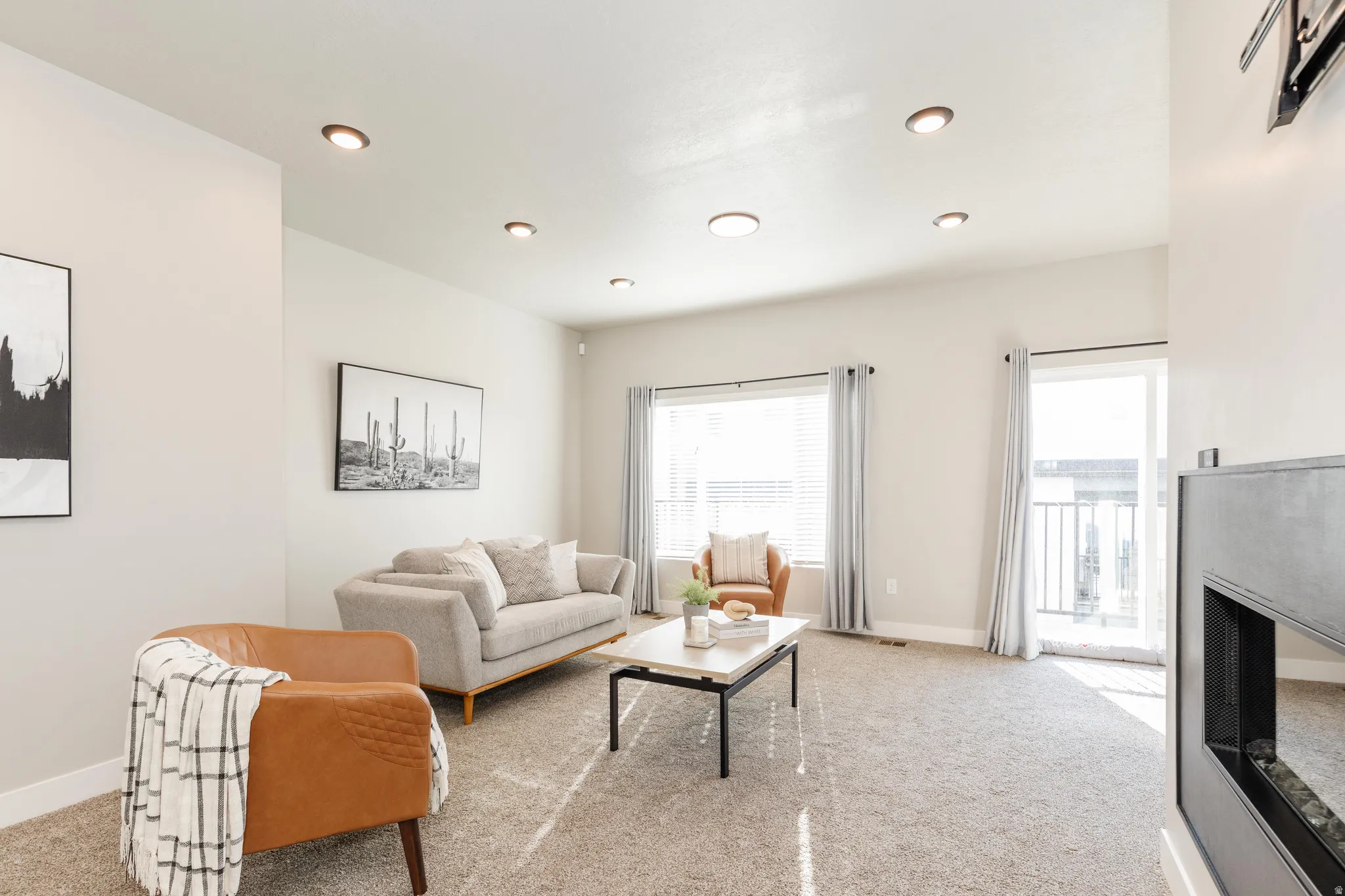 Spacious living area featuring light gray carpeting, recessed ceiling lighting, a fireplace with a dark gray surround, a large window with blinds, and a glass sliding door leading to an exterior balcony with a railing