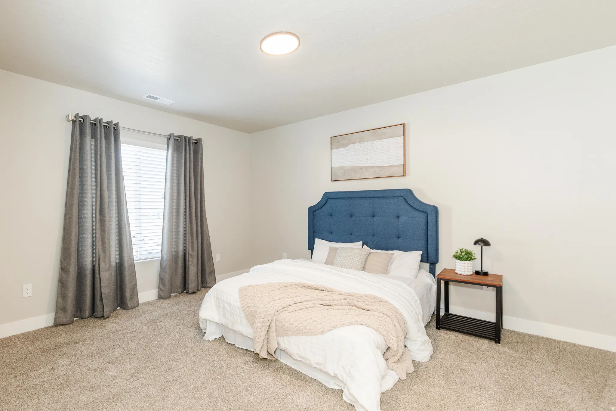 Spacious primary bedroom featuring a tufted upholstered headboard in blue, soft neutral bedding, carpet flooring, and abundant natural light from dual windows dressed in gray thermal curtains
