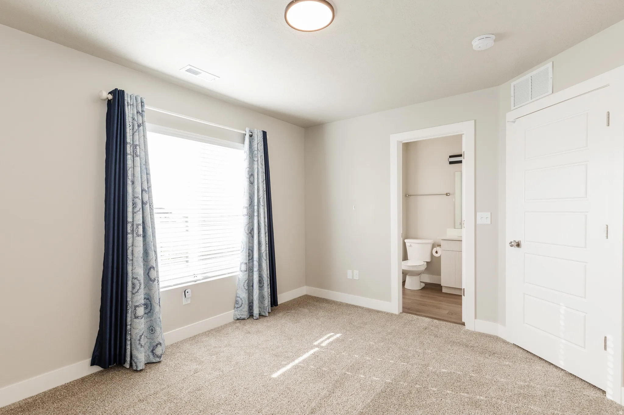 Carpeted room featuring a large window with blinds, recessed ceiling light, and an open doorway revealing a bathroom with a toilet and vanity
