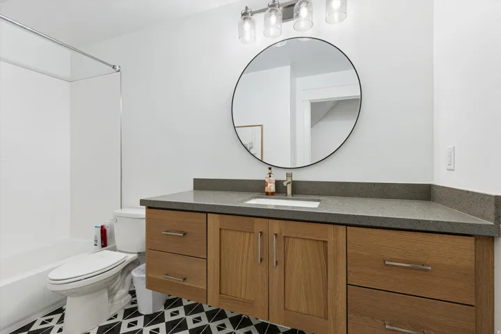 Bathroom featuring a wood-finish floating vanity with a gray countertop, an undermount sink, a circular mirror, and a black and white geometric tile floor