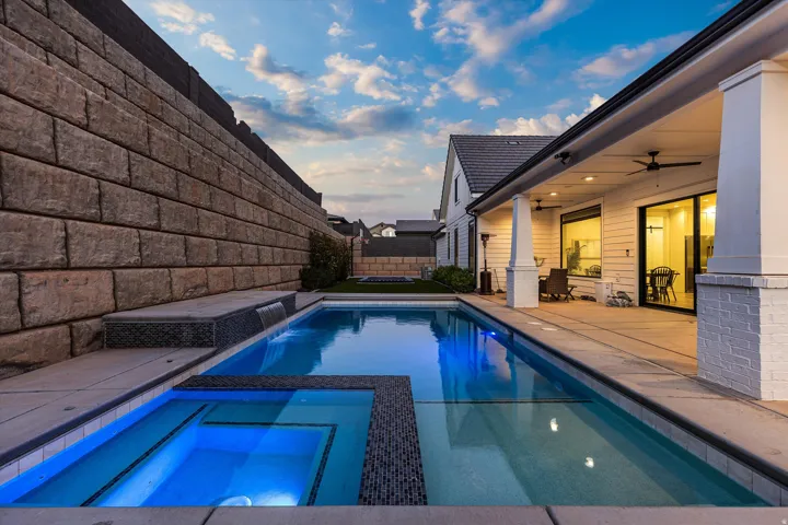 Expansive outdoor living area featuring a rectangular swimming pool with integrated spa, a tiered stone retaining wall, and a covered patio