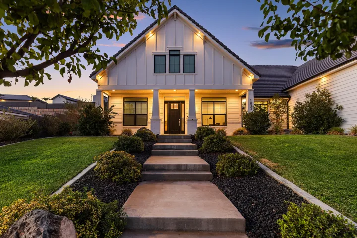 Contemporary farmhouse-style exterior featuring vertical board and batten siding, dark framed windows, a covered front porch with pillar supports, a concrete walkway with integrated steps, and a manicured lawn with shrubbery and dark rock landscaping