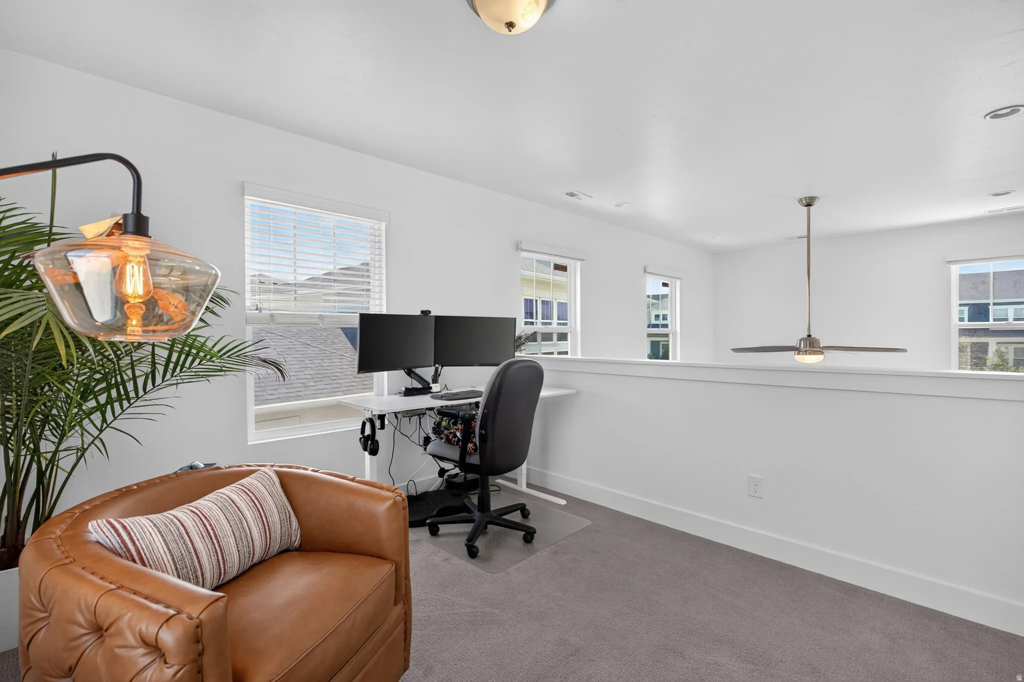Open loft space featuring light gray carpeting, multiple windows with blinds, a white half-wall, and a ceiling fan with integrated lighting