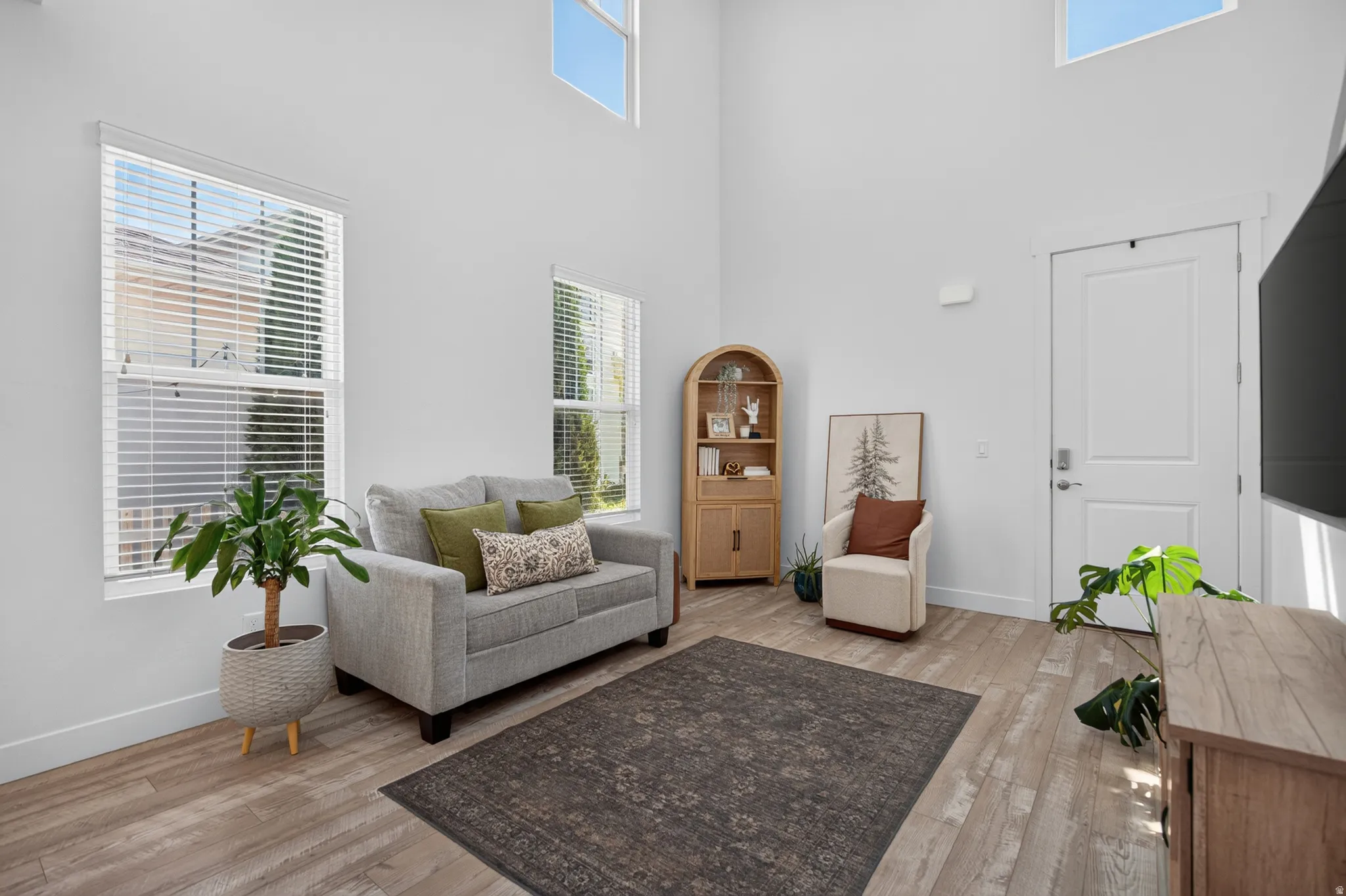 Living area featuring high ceilings, upper transom windows, two large windows with blinds, light wood-finish flooring, and a modern white door with brushed nickel hardware