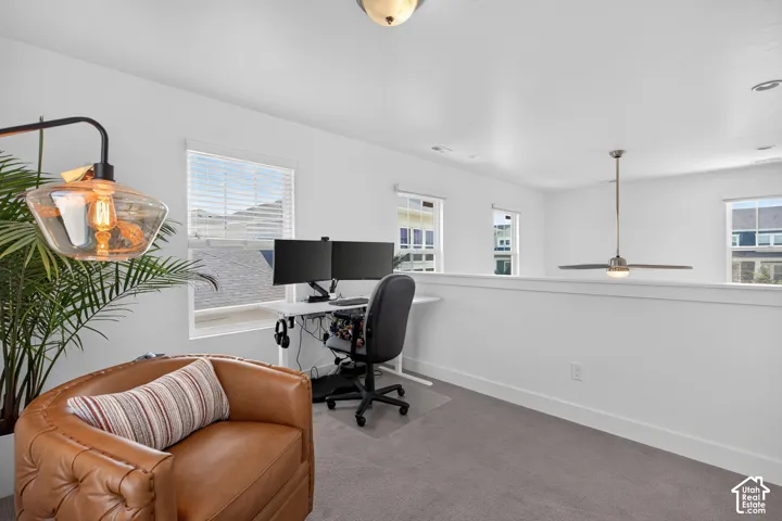 Open loft space featuring light gray carpeting, multiple windows with blinds, a white half-wall, and a ceiling fan with integrated lighting