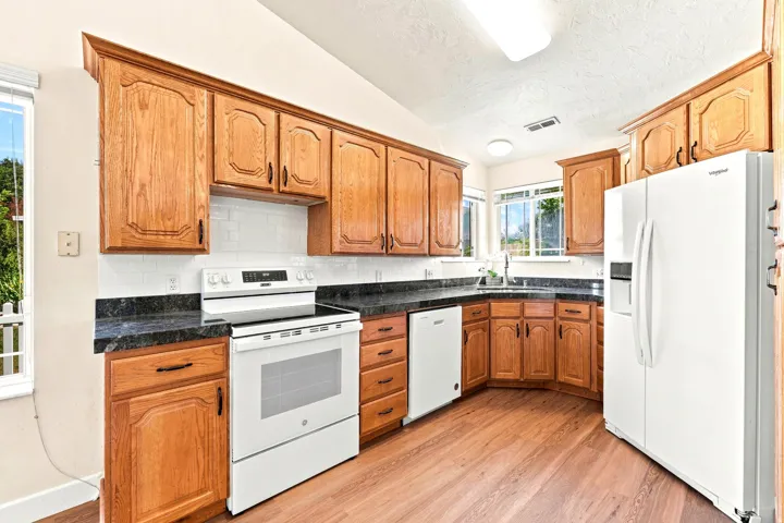 Kitchen featuring wood-finish flooring, wood cabinetry with dark hardware, dark countertops, white subway tile backsplash, and white appliances