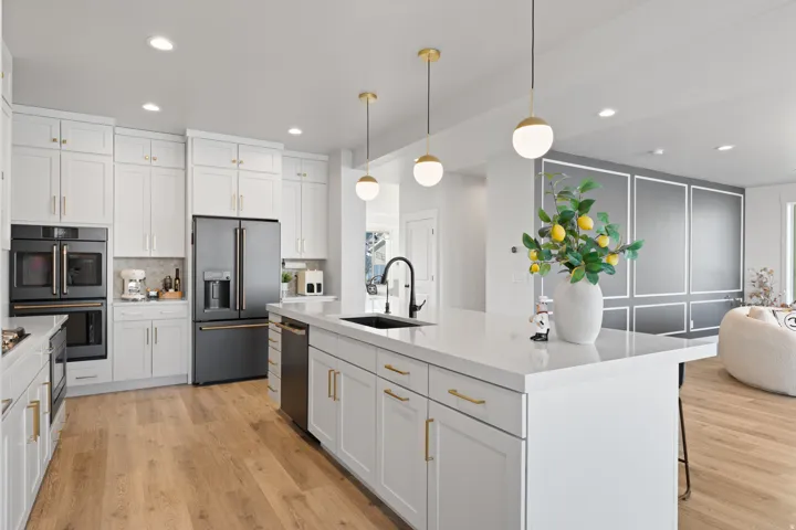 Modern kitchen featuring white cabinetry with gold-tone hardware, a large central island with a white countertop and integrated sink, stainless steel appliances, and light wood-finish flooring