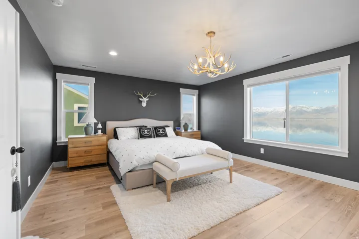 Bright bedroom featuring a large window with mountain and water scenery, light wood-finish flooring, recessed lighting, and a decorative antler chandelier
