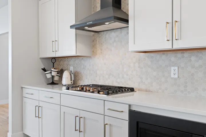 Modern kitchen featuring white shaker cabinetry, light-toned countertops, a gas cooktop with a black range hood, and a light-colored hexagonal tile backsplash