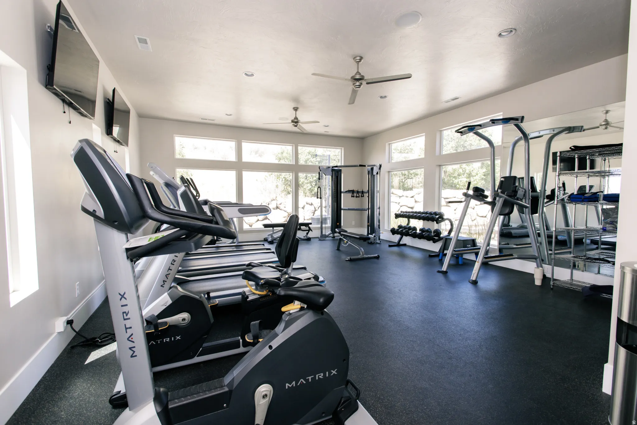 Dedicated fitness room featuring black rubber flooring, expansive windows, multiple recessed lights, and two ceiling fans