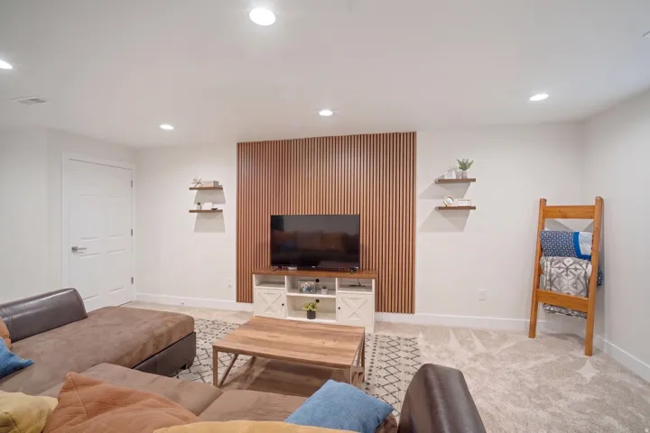 Finished basement recreation room featuring a wood-slat accent wall with integrated TV nook, floating shelves, recessed lighting, light wood-finish coffee table, and plush seating arrangement