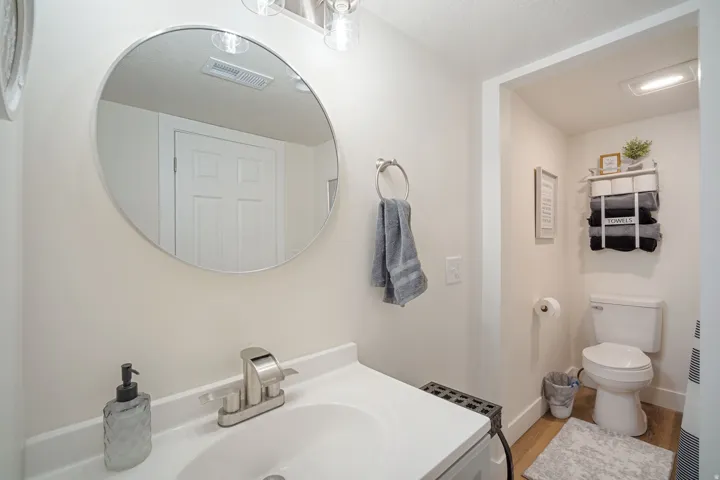Bathroom featuring a round wall-mounted mirror, brushed nickel faucet, a white vanity with integrated sink, and an alcove with a toilet