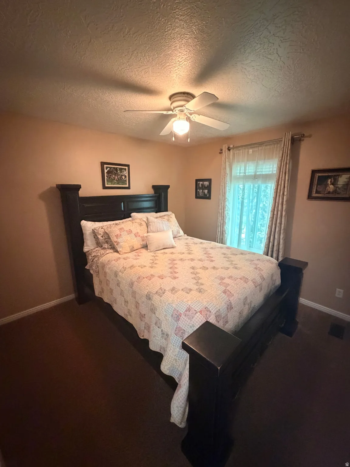 Bedroom featuring a ceiling fan with integrated lighting, textured ceilings, a window with sheer and patterned drapes, and dark carpeting