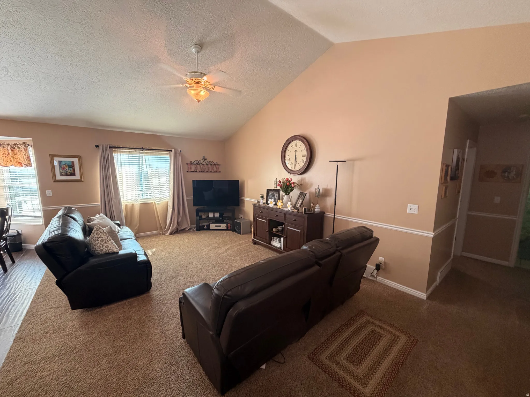 Spacious living area featuring a vaulted ceiling with a ceiling fan, carpet flooring, and a large window with blinds