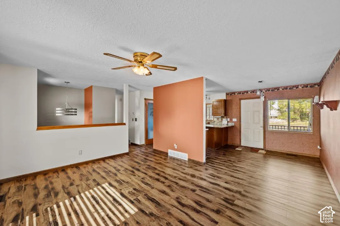 Wood-finish flooring throughout, ceiling fan with integrated lighting, kitchen area with wood cabinetry, white door with wood trim, and a window with horizontal blinds