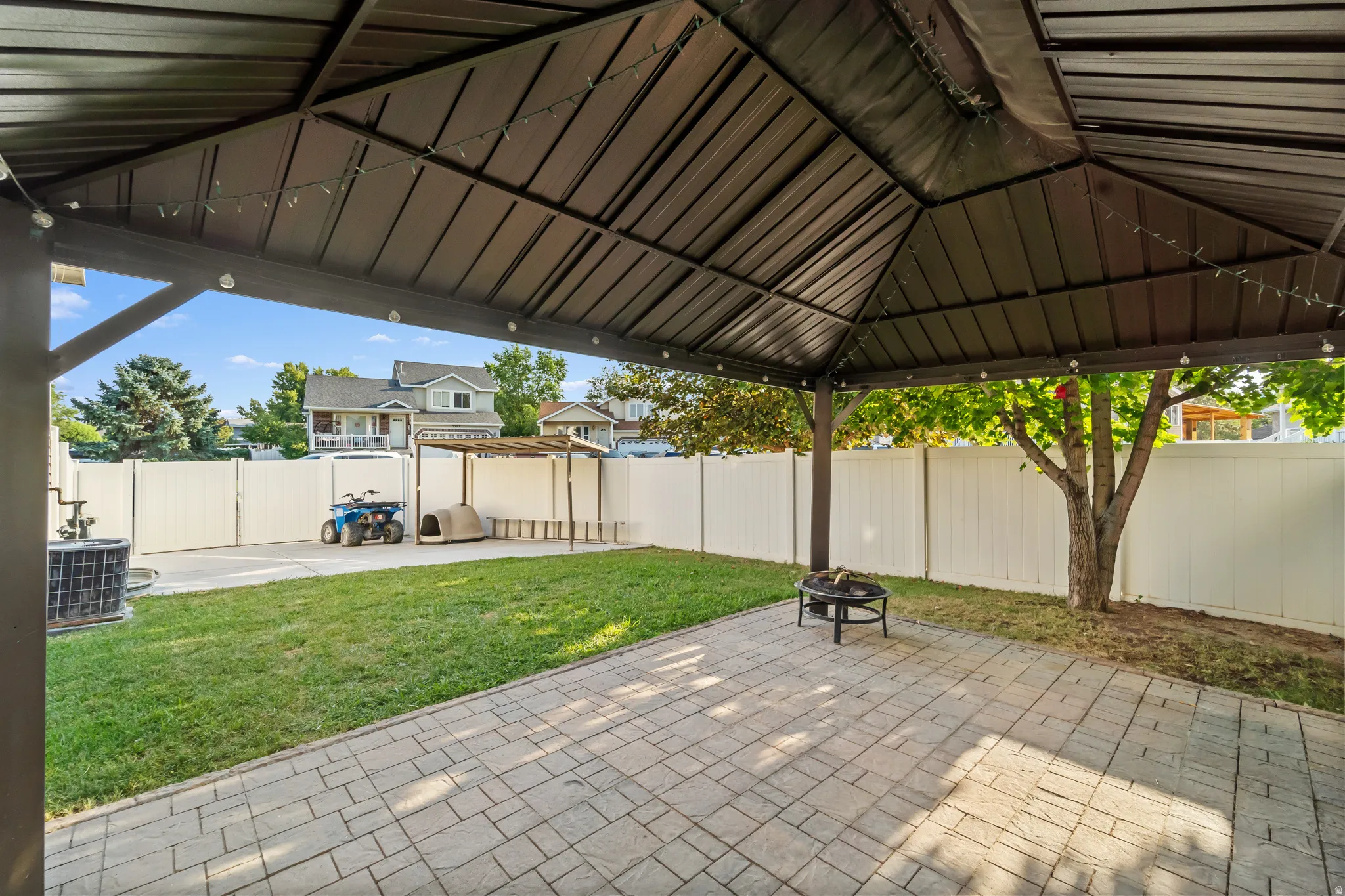 Outdoor living space featuring a dark-toned gazebo with string lights, a paver patio, a grass lawn, and a white privacy fence