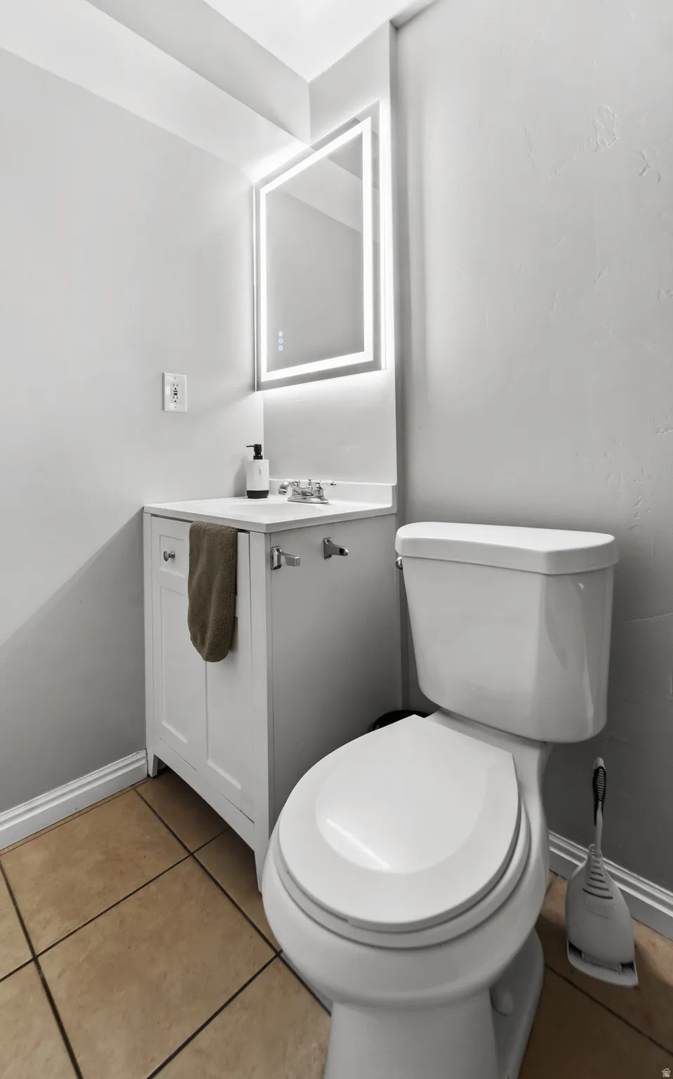 White vanity with integrated sink, chrome faucet, illuminated mirror, and neutral-toned tile flooring