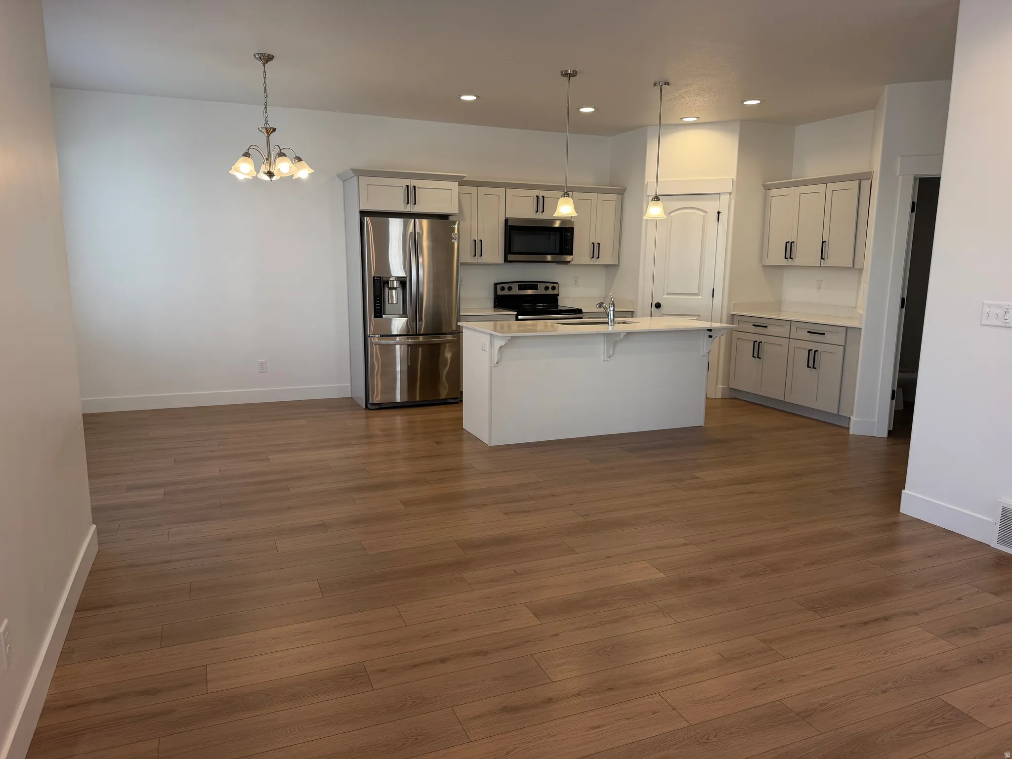Open-concept kitchen and dining area featuring white cabinetry, stainless steel appliances including a French-door refrigerator, a centered island with seating, wood-finish flooring throughout, recessed ceiling lighting, and pendant fixtures above the mai