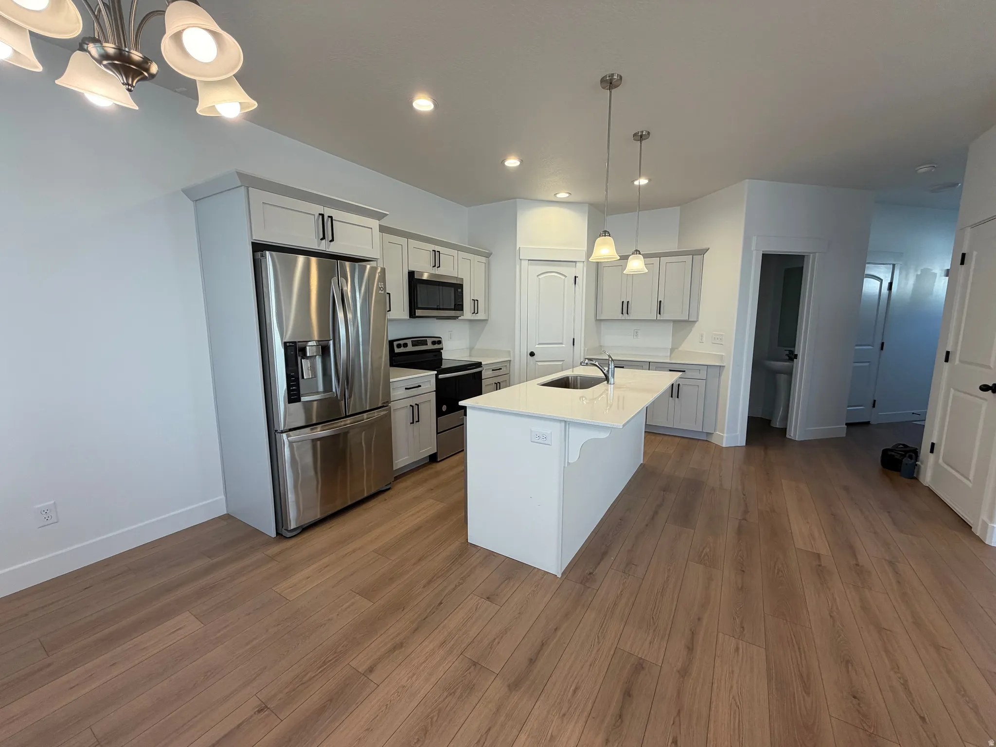 Contemporary kitchen featuring an island with an undermount sink, pendant lighting, stainless steel appliances, light wood-finish flooring, and white cabinetry