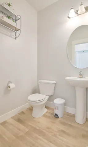 Powder room featuring light wood-finish flooring, a white pedestal sink, a round wall-mounted mirror, and a two-bulb vanity light fixture