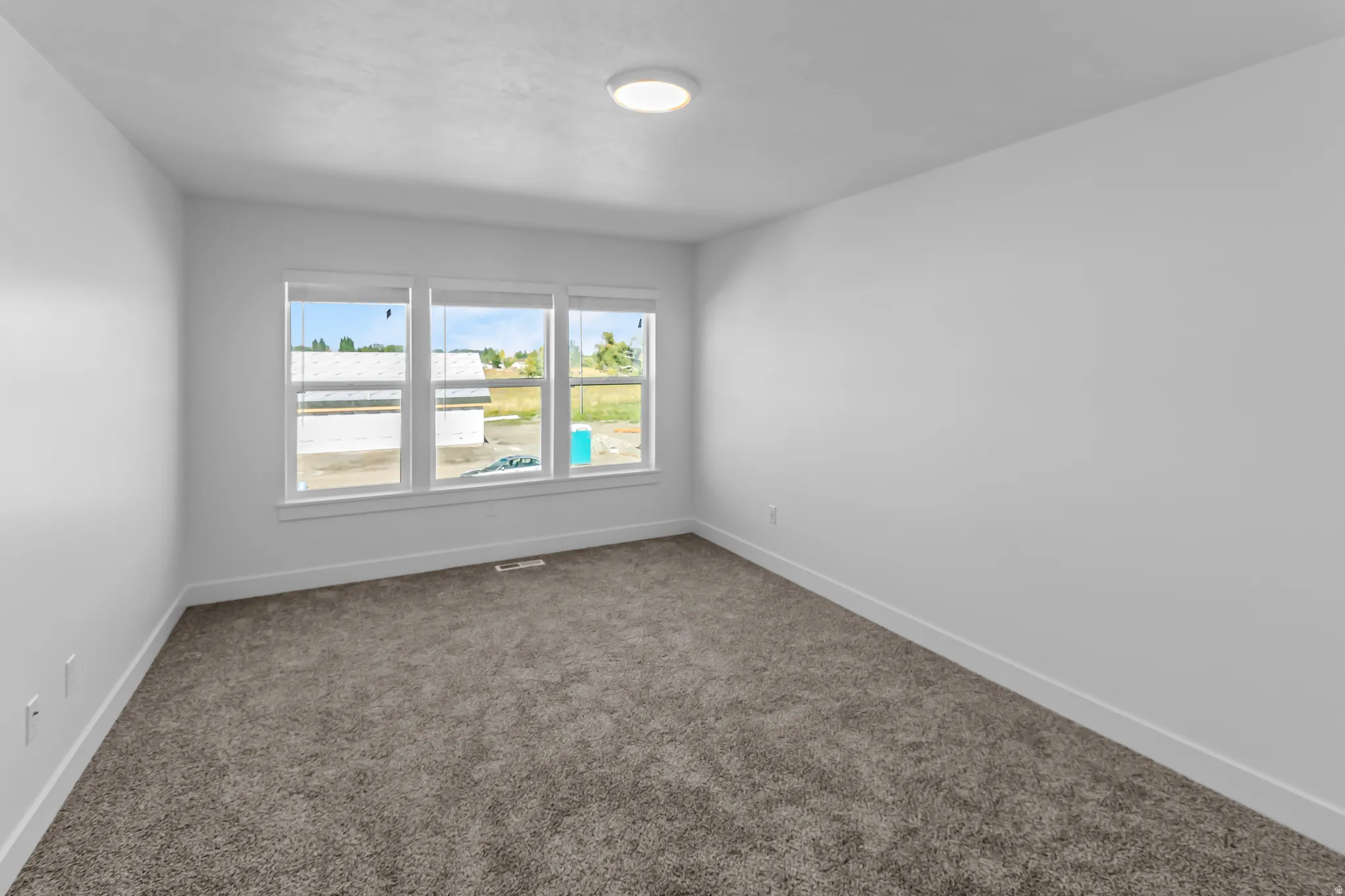 Light-filled room featuring three large windows, plush gray carpeting, white walls, white trim, and a flush-mount ceiling light fixture