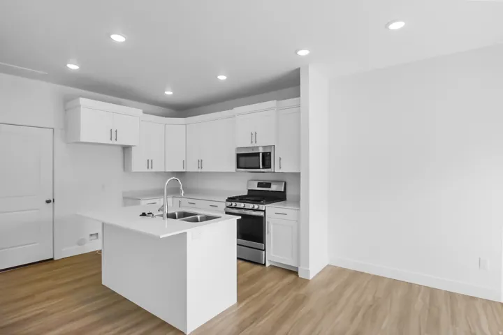 Kitchen featuring white shaker-style cabinetry, a central island with a double basin sink and gooseneck faucet, stainless steel appliances, recessed ceiling lighting, and wood-finish flooring