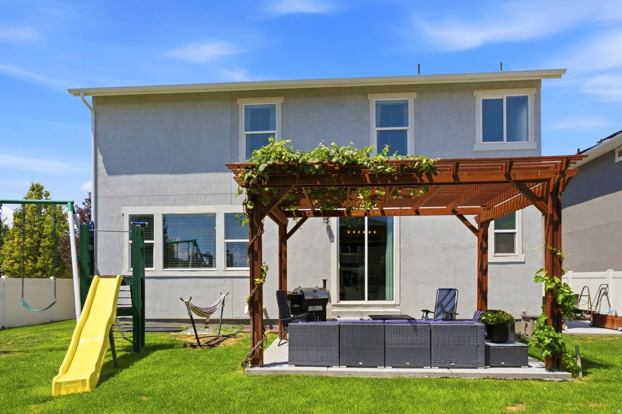 Expansive backyard featuring a lush lawn, a pergola-covered patio with concrete flooring, a swing set with a slide, and a stucco exterior