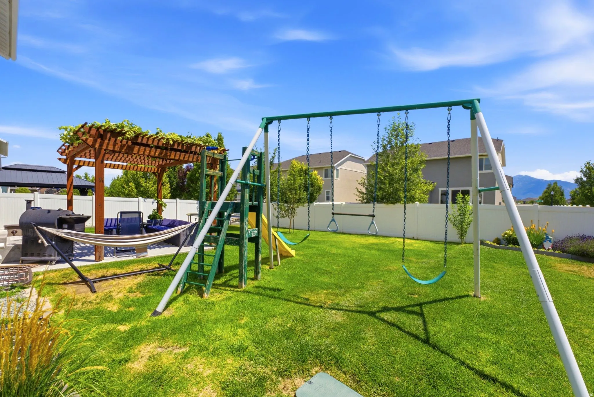 Expansive backyard featuring a lush lawn, a wooden pergola with mature vines, a concrete patio area, a white privacy fence, and distant mountain scenery