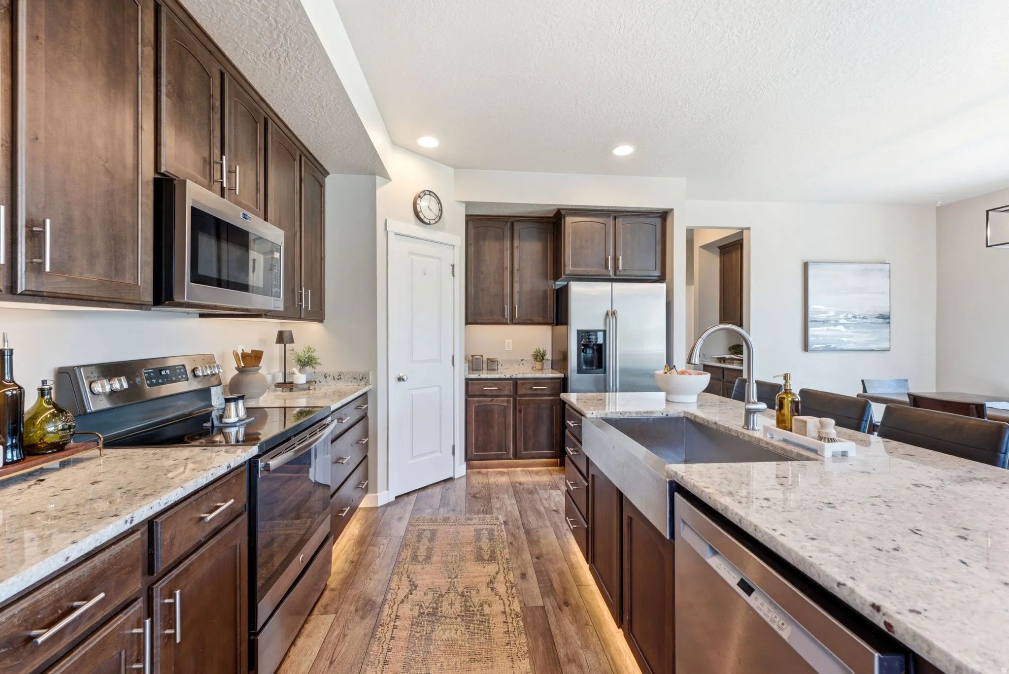 Open-concept kitchen featuring dark wood cabinetry, light granite countertops, stainless steel appliances, a farmhouse sink, and wood-finish flooring