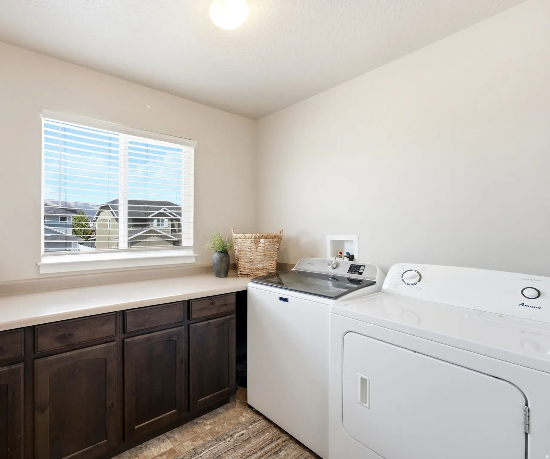Utility room featuring a window with horizontal blinds, extensive countertop space, dark wood-finish cabinetry with recessed panels, and durable wood-look flooring