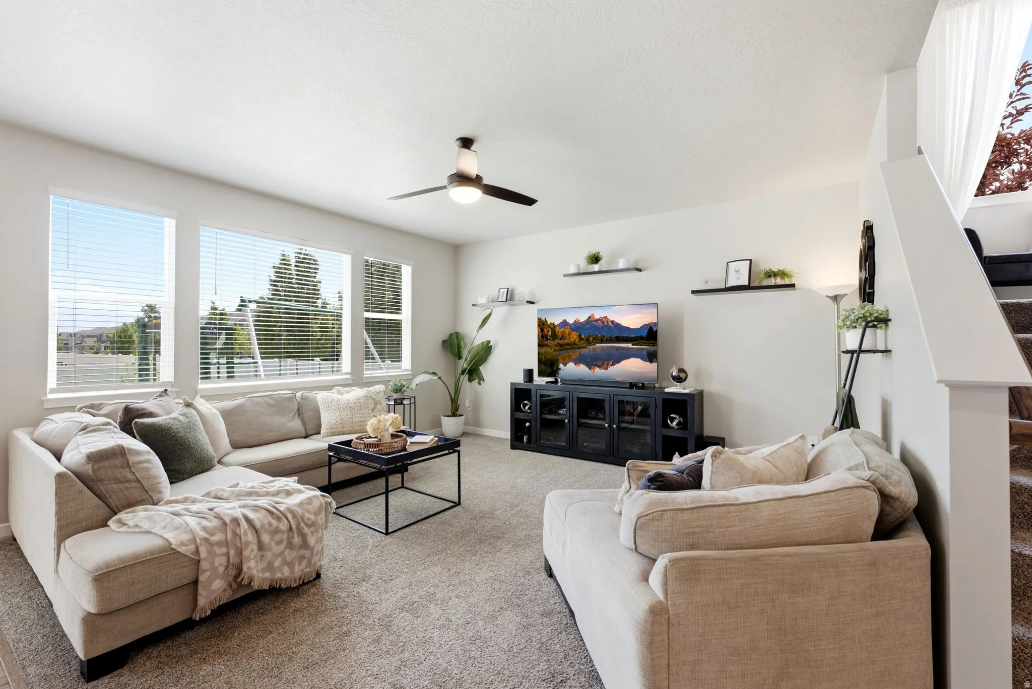 Living area featuring multiple large windows, light-toned carpeting, a ceiling fan with integrated lighting, and a staircase with a white railing