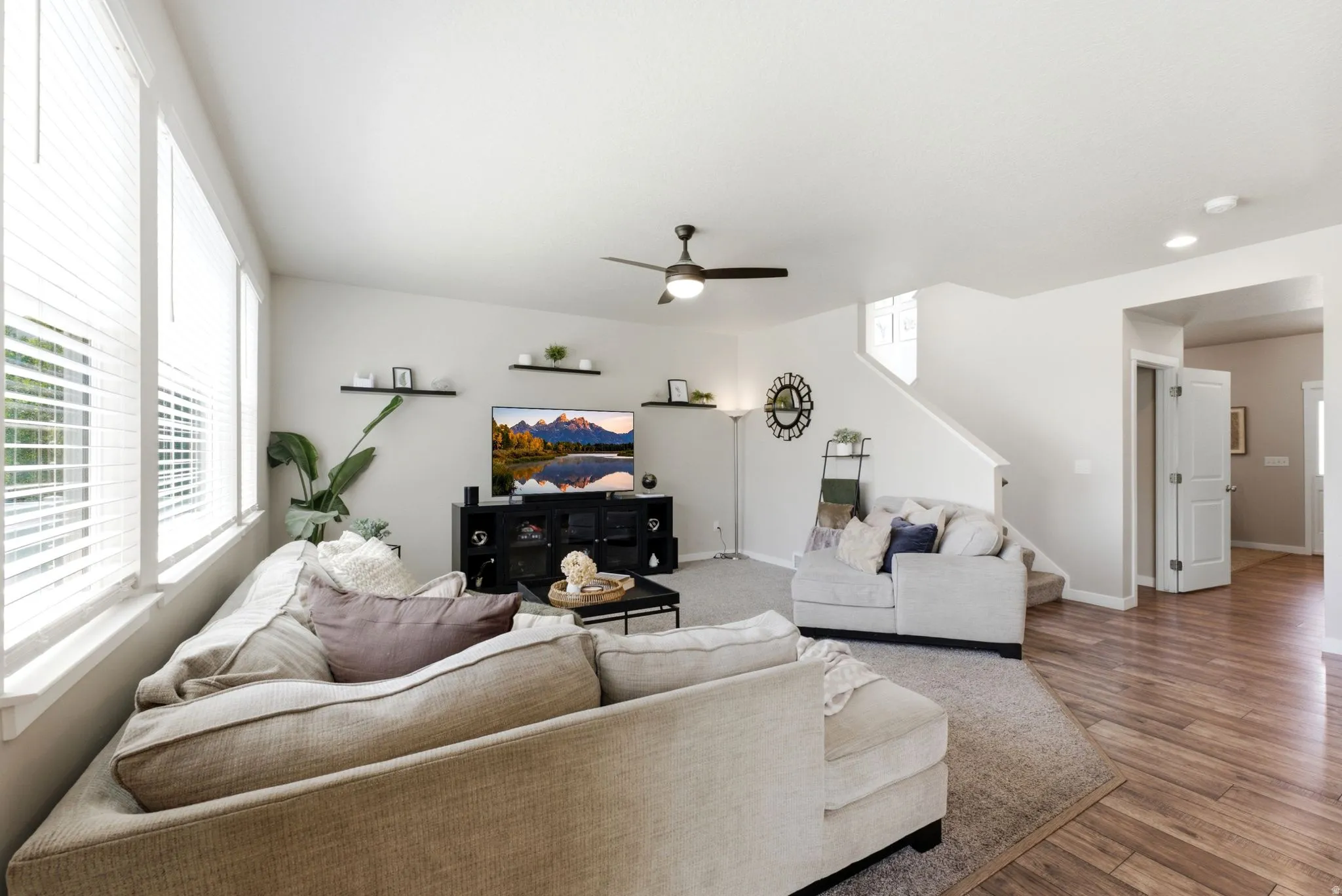 Open-concept living space featuring wood-finish flooring, a contemporary ceiling fan, multiple large windows with blinds, and a staircase with a carpet runner