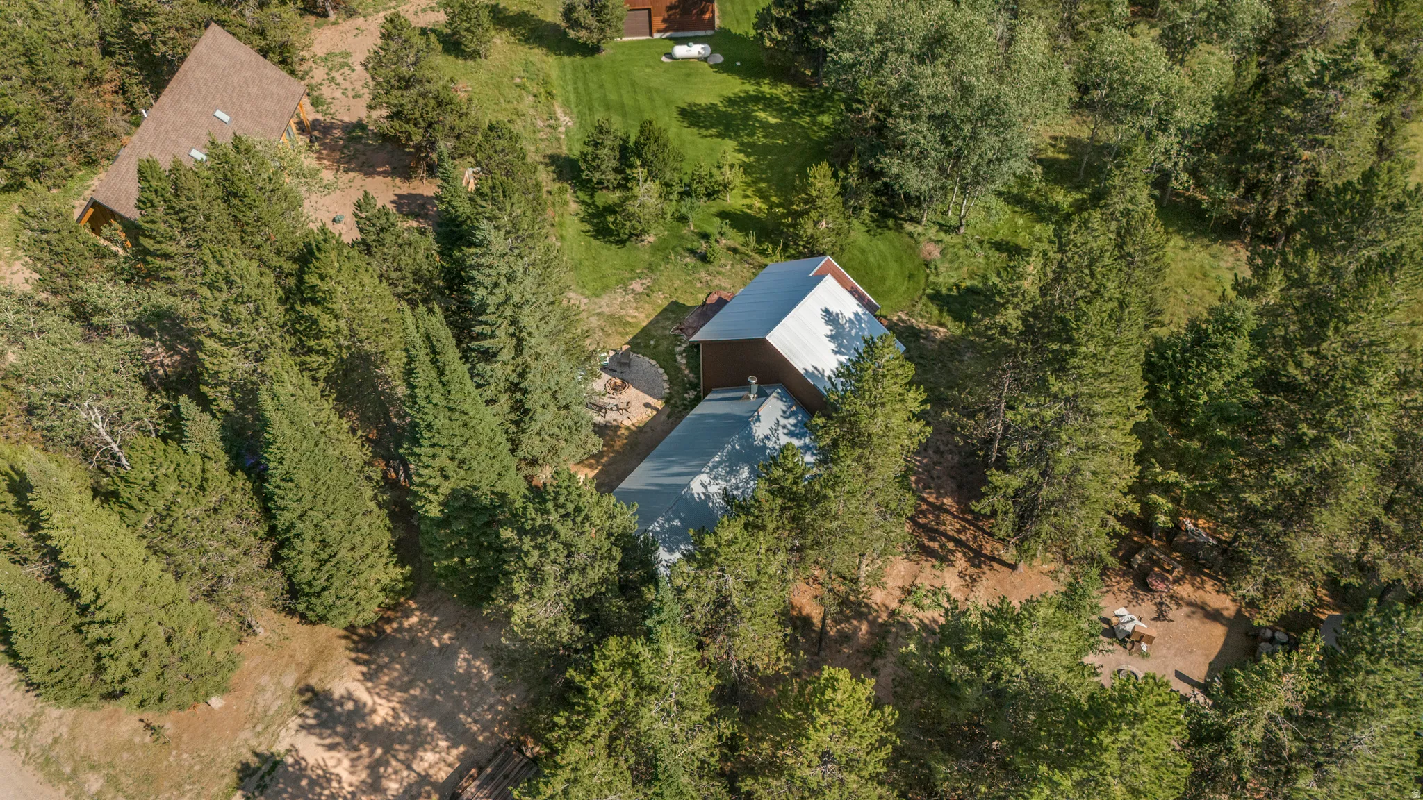 Aerial view of a property surrounded by mature trees, featuring a main residence with a metal roof and a second structure with a shingled roof