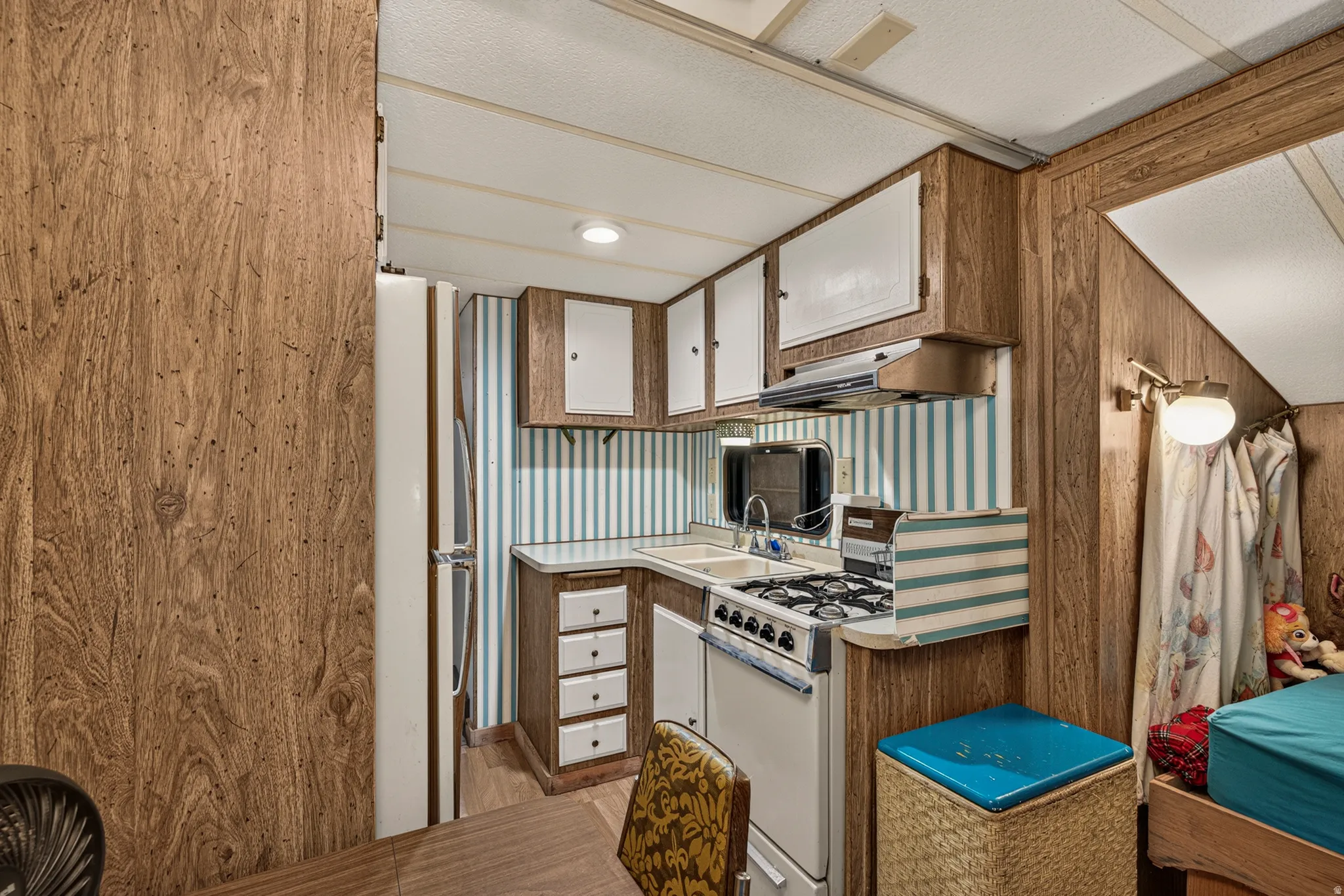 Compact kitchen featuring wood-finish paneling, overhead cabinetry with white doors, a gas range with an exhaust hood, and a built-in sink with a mirrored backsplash