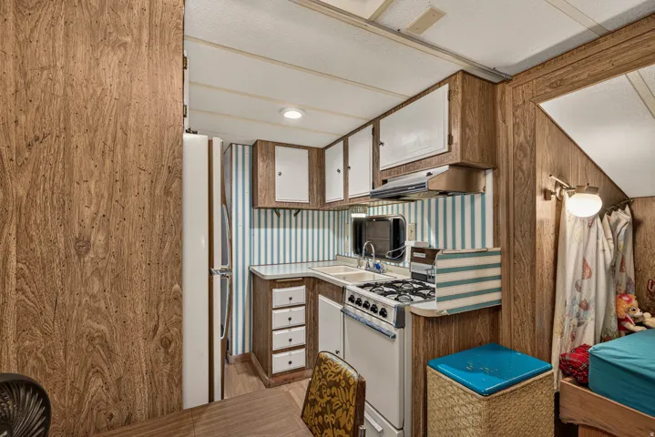 Compact kitchen featuring wood-finish paneling, overhead cabinetry with white doors, a gas range with an exhaust hood, and a built-in sink with a mirrored backsplash