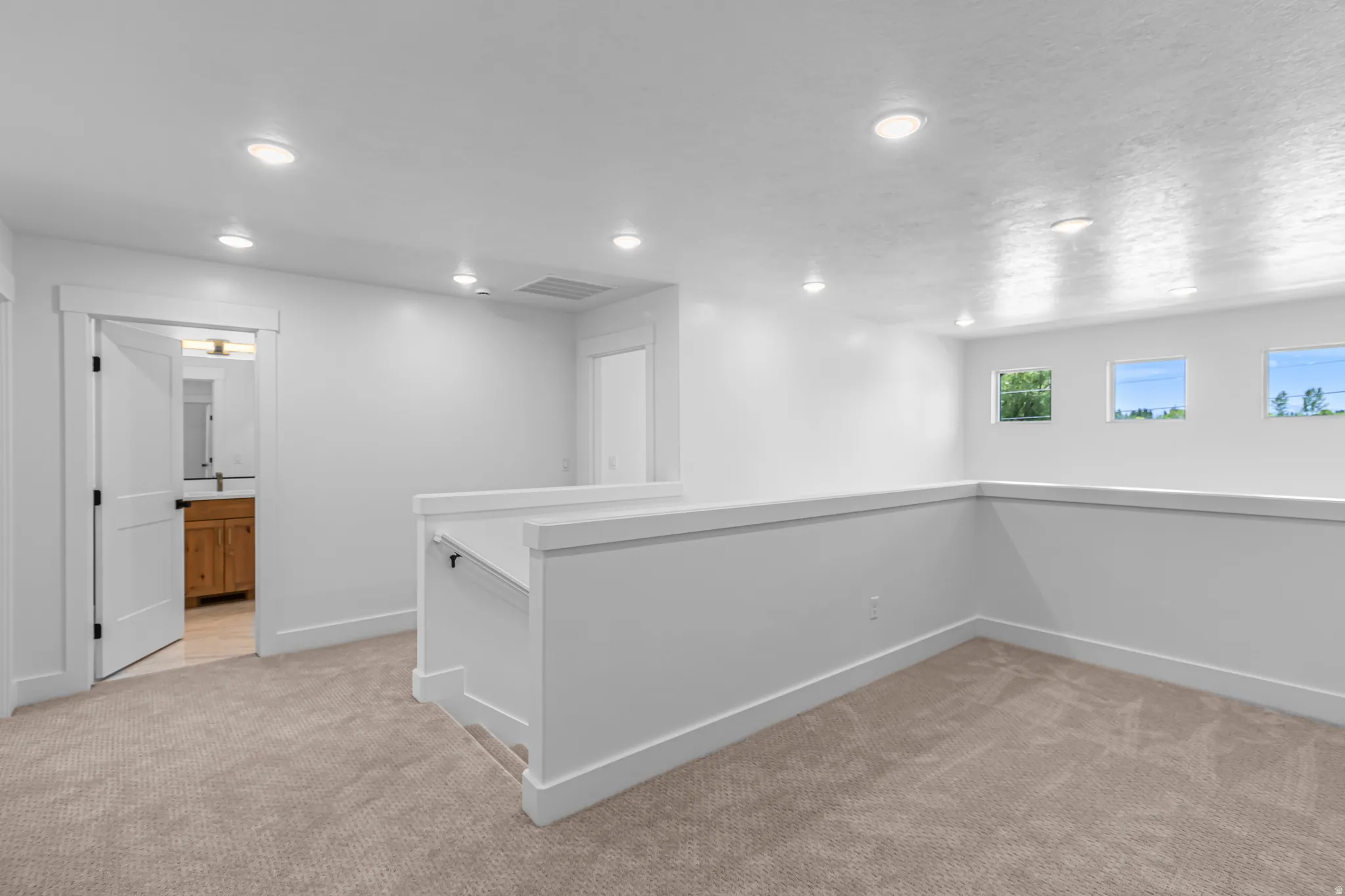 Upper-level landing featuring recessed lighting, plush carpeting, multiple windows, a white painted railing, and an adjacent bathroom with a wood-finish vanity