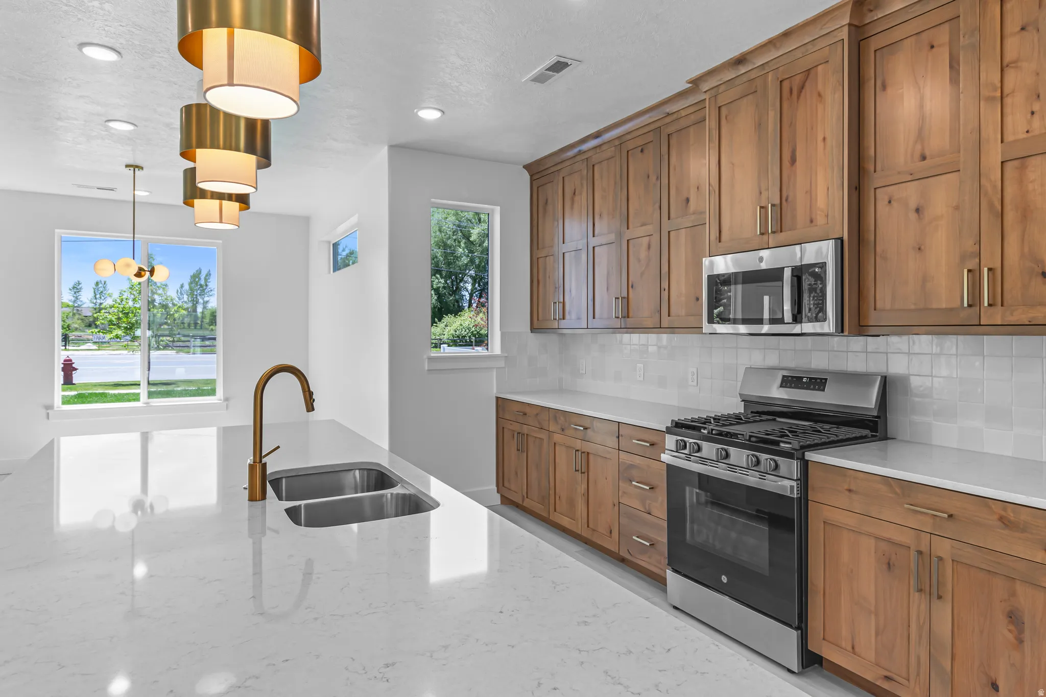 Kitchen featuring a large island with an undermount double-basin sink and brass-finish faucet, light-toned countertops, wood cabinetry with gold-finish hardware, stainless steel appliances, and a white subway tile backsplash